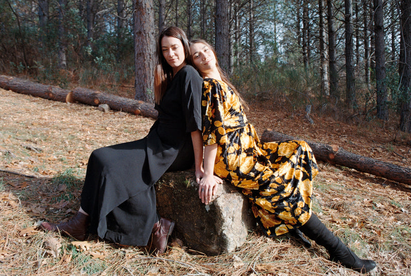 2 WOMEN SITTING ON A ROCK WEARING A SLOW FASHIO WRAP DRESS WITH VELVET FABRIC AND A FLORAL PRINT ON ONE AND BLACK LINEN COTTON ON THE OTHER