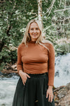 Woman in a brown top and black skirt standing in front of a forest with a waterfall.