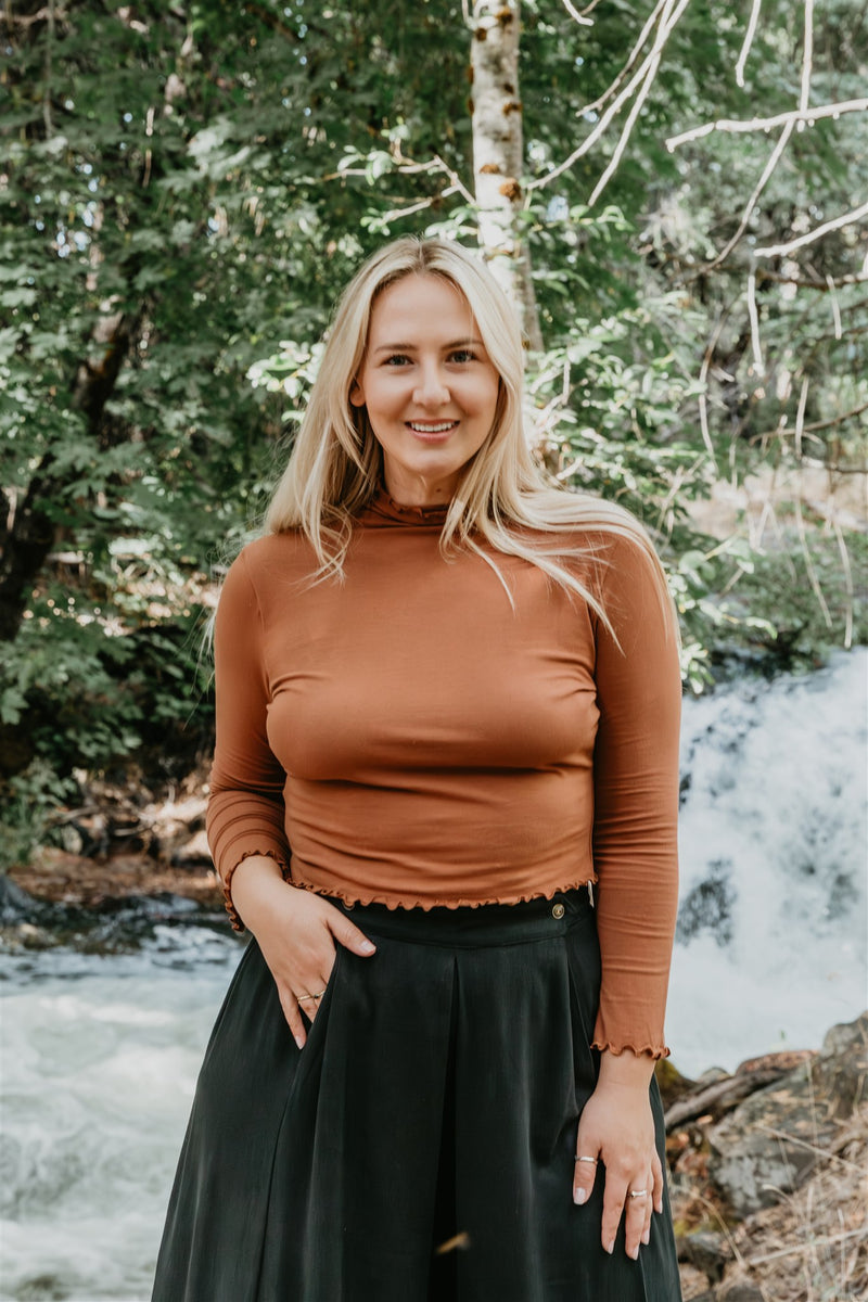 Woman in a brown top and black skirt standing in front of a forest with a waterfall.