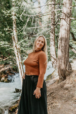Woman standing in a forest with a stream in the background waring a brown shirt and black wide leg pants