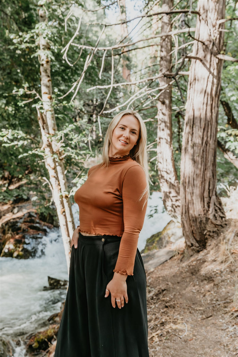 Woman standing in a forest with a stream in the background waring a brown shirt and black wide leg pants